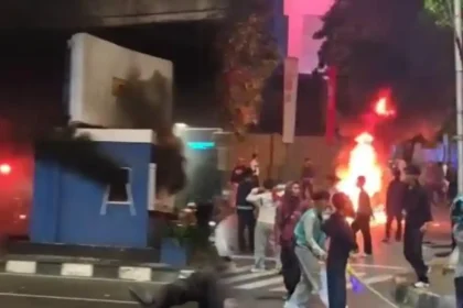 Nighttime street protest with a large fire and smoke in the background; people move around debris.