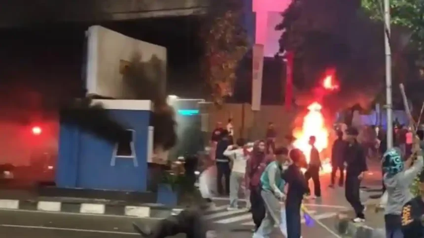 Nighttime street protest with a large fire and smoke in the background; people move around debris.