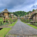 Stone-paved central path through a Balinese temple complex, flanked by carved shrines and tropical plants under a cloudy sky on both sides.