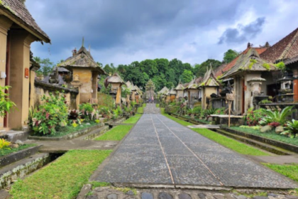 Stone-paved central path through a Balinese temple complex, flanked by carved shrines and tropical plants under a cloudy sky on both sides.