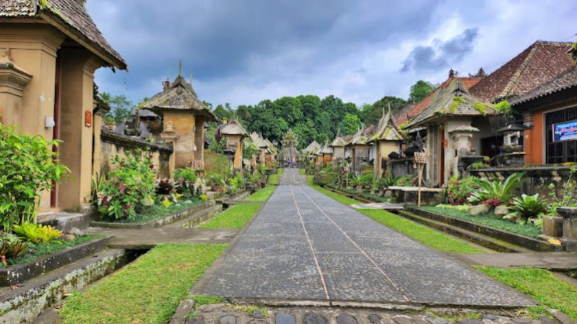 Stone-paved central path through a Balinese temple complex, flanked by carved shrines and tropical plants under a cloudy sky on both sides.