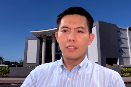Man speaking during a video call with a modern campus-style building in the background, wearing a light blue striped button-down shirt