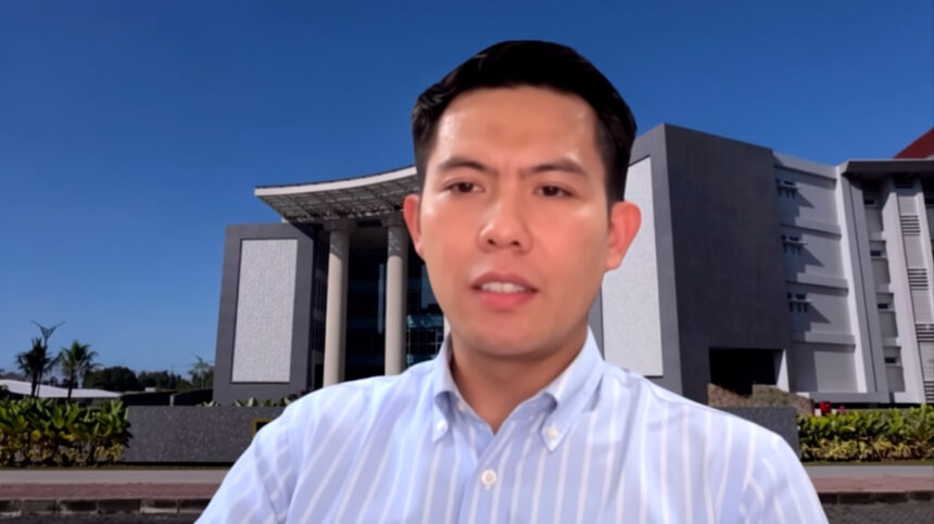 Man speaking during a video call with a modern campus-style building in the background, wearing a light blue striped button-down shirt