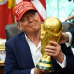 Man in a dark suit wearing a red cap with white text holds a large gold trophy in an office setting, smiling with eyes closed as another person points toward the trophy.