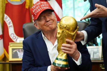 Man in a dark suit wearing a red cap with white text holds a large gold trophy in an office setting, smiling with eyes closed as another person points toward the trophy.