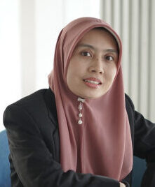 Woman in a pink hijab and black blazer sits at a desk in an office, looking at the camera.