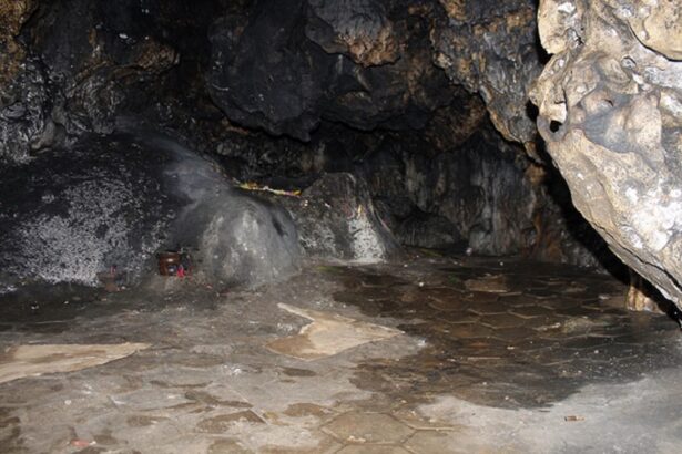 Dark cave interior with a wet, uneven stone floor and rugged rock walls. A shallow pool of water is visible on the right side.
