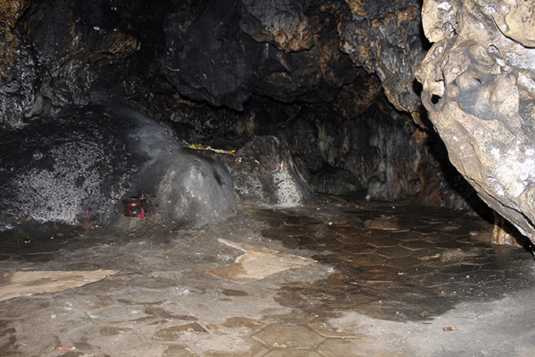 Dark cave interior with a wet, uneven stone floor and rugged rock walls. A shallow pool of water is visible on the right side.