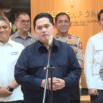 Central man speaks at a microphone during a briefing, surrounded by uniformed officials in a formal setting with a wood-paneled backdrop.