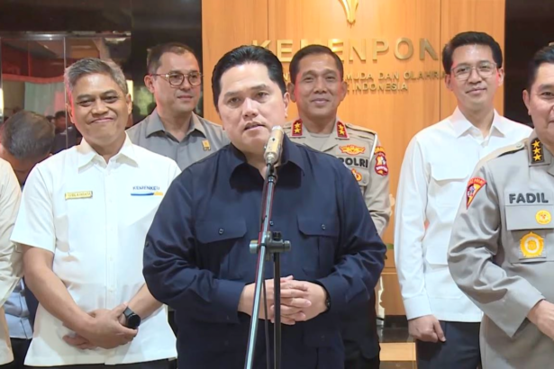 Central man speaks at a microphone during a briefing, surrounded by uniformed officials in a formal setting with a wood-paneled backdrop.