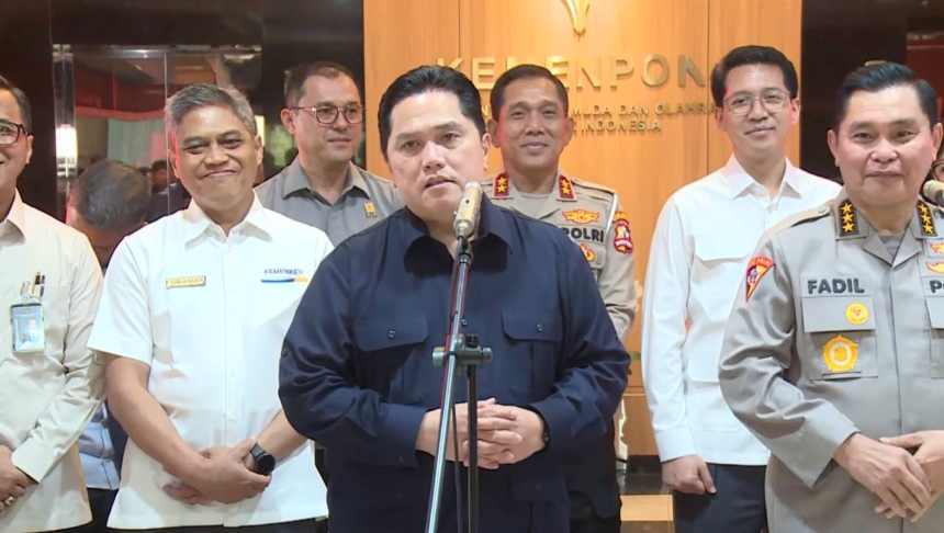 Central man speaks at a microphone during a briefing, surrounded by uniformed officials in a formal setting with a wood-paneled backdrop.
