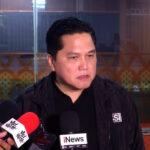 Man in a black jacket speaks to reporters with multiple microphones in front, indoor press area in background