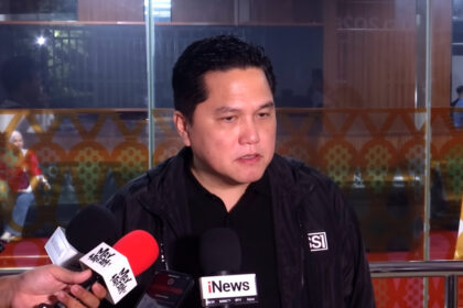 Man in a black jacket speaks to reporters with multiple microphones in front, indoor press area in background