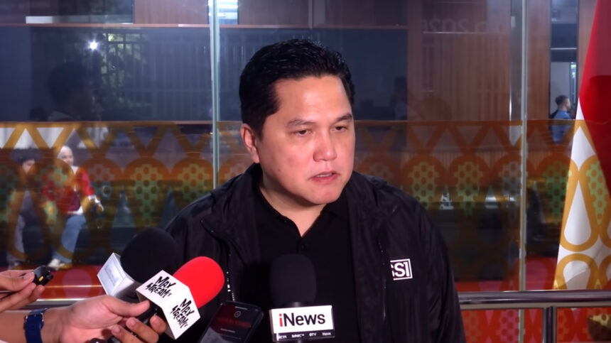 Man in a black jacket speaks to reporters with multiple microphones in front, indoor press area in background