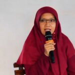 Woman in a maroon hijab and glasses speaks into a handheld microphone while seated against a neutral background