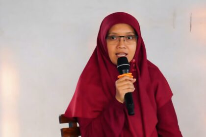 Woman in a maroon hijab and glasses speaks into a handheld microphone while seated against a neutral background