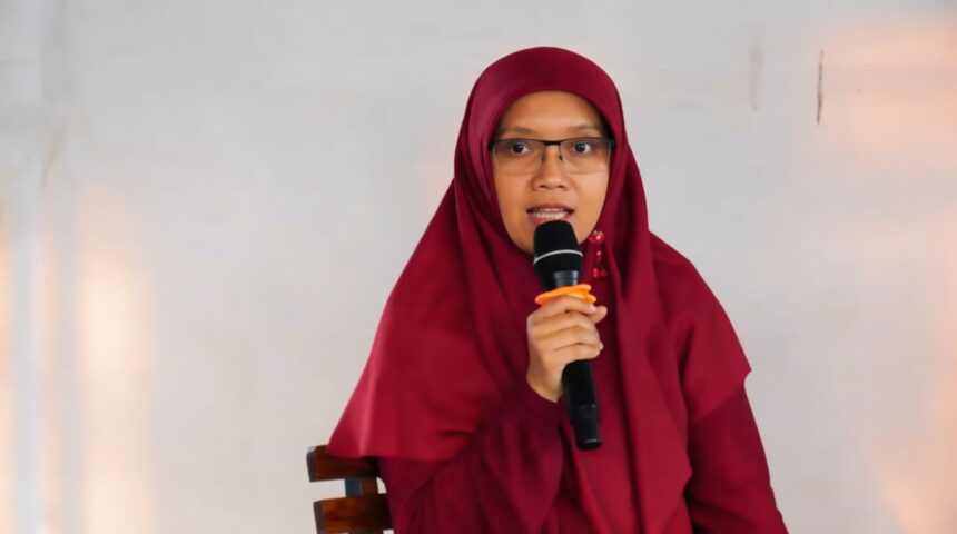 Woman in a maroon hijab and glasses speaks into a handheld microphone while seated against a neutral background
