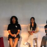 Panel of four women seated on stage; leftmost in maroon hijab speaks into a microphone against a neutral backdrop.