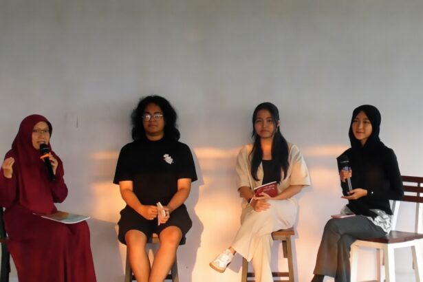 Panel of four women seated on stage; leftmost in maroon hijab speaks into a microphone against a neutral backdrop.