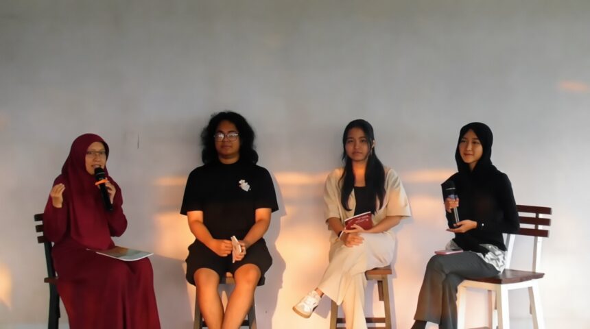 Panel of four women seated on stage; leftmost in maroon hijab speaks into a microphone against a neutral backdrop.