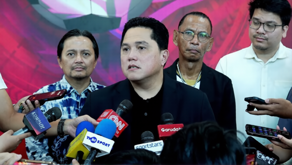 Spokesperson in a black suit speaks at a press conference, reporters surround him with multiple microphones and phones nearby.