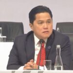 Man in a dark suit and red tie sits at a panel table, speaking into a microphone during a conference. (informative)
