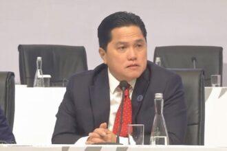 Man in a dark suit and red tie sits at a panel table, speaking into a microphone during a conference. (informative)
