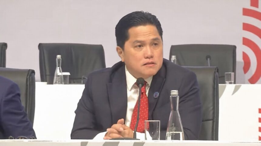 Man in a dark suit and red tie sits at a panel table, speaking into a microphone during a conference. (informative)