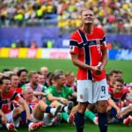 Norway national team players sit on the pitch in red jerseys while a teammate stands holding a drum, smiling at cheering fans in the stadium light.