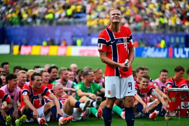 Norway national team players sit on the pitch in red jerseys while a teammate stands holding a drum, smiling at cheering fans in the stadium light.