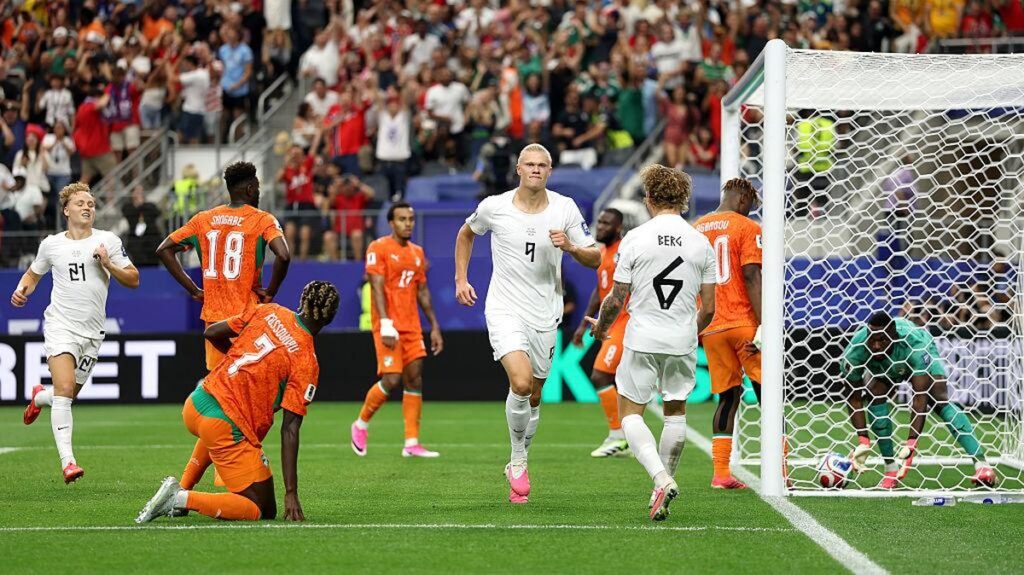 Erling Haaland vs Pantai Gading by Getty Images - Mabur.co Soccer match scene with white team players celebrating near the goal as the ball sits by the net; orange team players nearby and a goalkeeper in green reacts near the goal line.