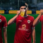 Three men in red soccer jerseys drink beer mugs during a break on a green field with sponsor banners nearby.