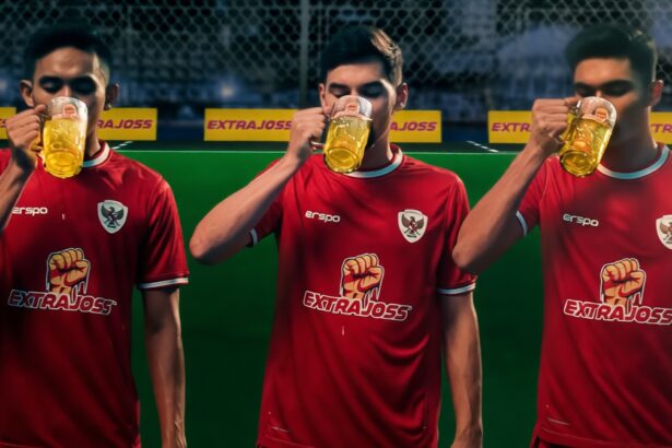 Three men in red soccer jerseys drink beer mugs during a break on a green field with sponsor banners nearby.