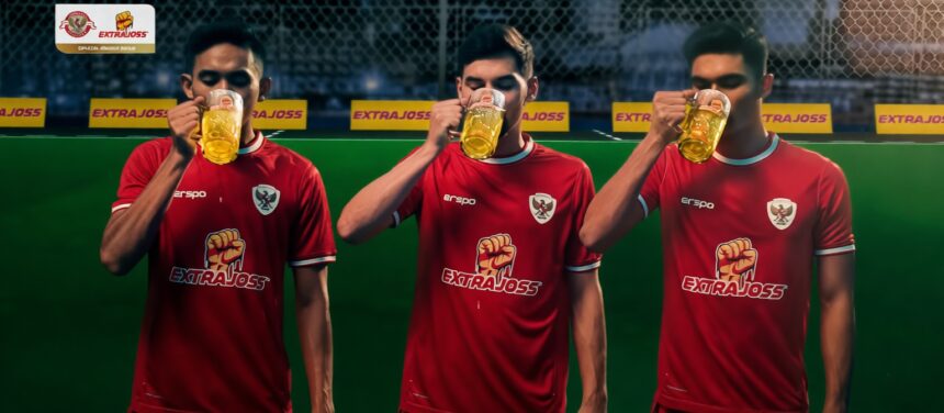Three men in red soccer jerseys drink beer mugs during a break on a green field with sponsor banners nearby.