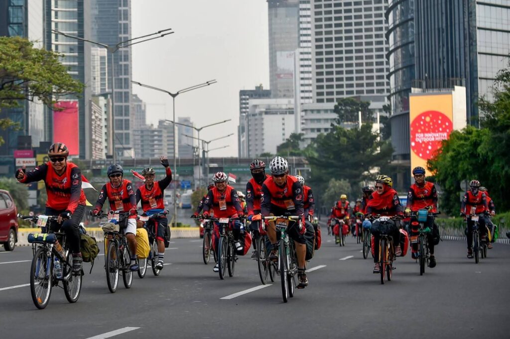 FB_IMG_1783215985294 - Mabur.co Group of cyclists in red jerseys riding down a city street with tall buildings in the background, during a parade or race.