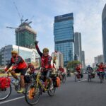 Group of cyclists wearing red vests ride on a city street during a road race, with tall modern buildings in the background.