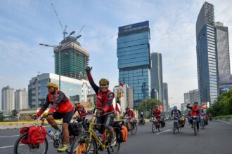 Group of cyclists wearing red vests ride on a city street during a road race, with tall modern buildings in the background.