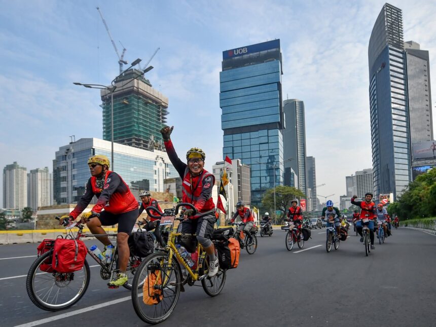 FB_IMG_1783216115399 - Mabur.co Group of cyclists wearing red vests ride on a city street during a road race, with tall modern buildings in the background.