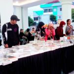 Long table covered with books at an outdoor book fair, with people browsing and chatting nearby.