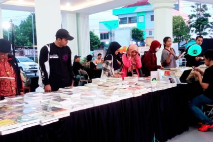 Long table covered with books at an outdoor book fair, with people browsing and chatting nearby.