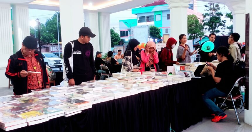 Long table covered with books at an outdoor book fair, with people browsing and chatting nearby.