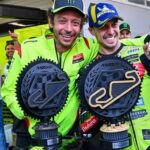 Two racers in neon green suits celebrate with oversized circular trophies in a pit area, posing and smiling for the camera.