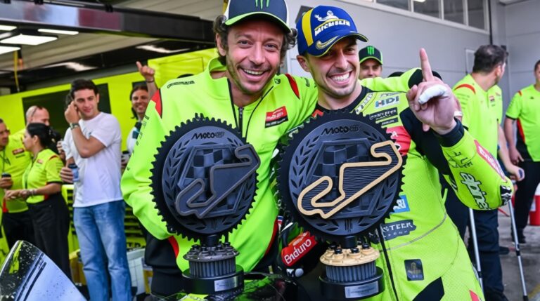 Fabio Diggia V Rossi by Motogp com - Mabur.co Two racers in neon green suits celebrate with oversized circular trophies in a pit area, posing and smiling for the camera.
