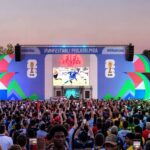 Crowd of fans at an outdoor FIFA Fan Fest with a large colorful stage, big screen showing a soccer match, and waving flags at sunset.