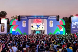 Crowd of fans at an outdoor FIFA Fan Fest with a large colorful stage, big screen showing a soccer match, and waving flags at sunset.