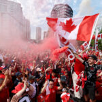 Crowd of Canadians celebrating with red clothing and waving Canadian flags amid pink smoke and city buildings in the background.