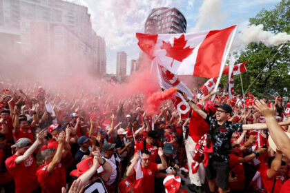 Crowd of Canadians celebrating with red clothing and waving Canadian flags amid pink smoke and city buildings in the background.