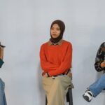Three women sit on stools in a panel discussion; the leftmost speaker holds a microphone and talks, while the other two listen nearby.