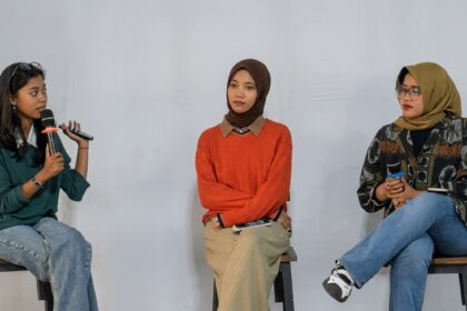 Three women sit on stools in a panel discussion; the leftmost speaker holds a microphone and talks, while the other two listen nearby.
