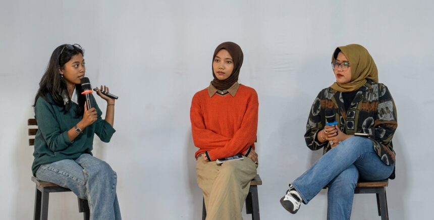 Three women sit on stools in a panel discussion; the leftmost speaker holds a microphone and talks, while the other two listen nearby.